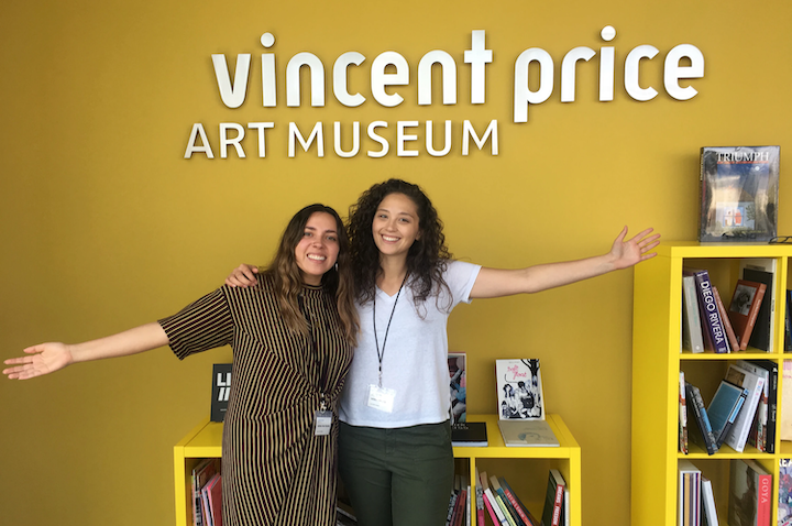 Two women students welcoming visitors in front of the Vincent Proce Art Museum sign.