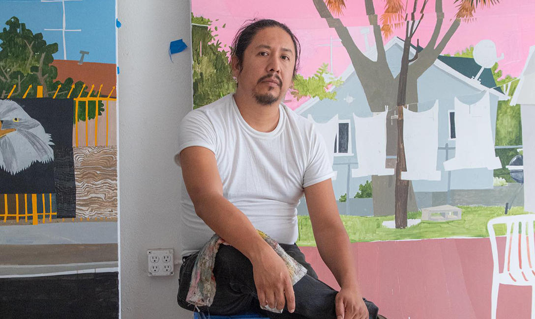 Man with dark hair and facial hair seated facing the camera in front of paintings of houses that include trees, a fence, and lawn chair and clotheslines.